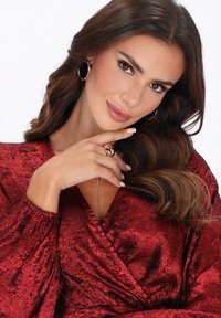 Frau mit langen braunen Haaren, die goldene Ohrringe, einen Ring und ein rotes Samtwickelkleid trägt, posiert mit der Hand nahe dem Kinn und lächelt sanft.