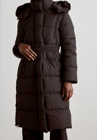 Černý péřový kabát s páskem v pase, vysokým límcem a kapucí s kožešinovým lemem, prošívaný design a dlouhé rukávy.