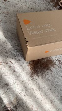 Barna kartondoboz nyomtatott szöveggel és Zalando logóval, egy világos színű, texturált szőrmés felületen pihen, amely sötét foltokkal díszített.