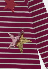 Fioletowo-białe w paseczki materiał z długimi rękawami, z dwoma odwracalnymi motywami muszelek z cekinów, jednym w kolorze różowym i złotym, a drugim czerwonym.