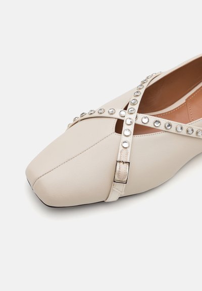 Chaussure plate beige avec un bout pointu, design à lanières croisées avec des clous argentés et un détail de boucle. Matériau synthétique lisse.