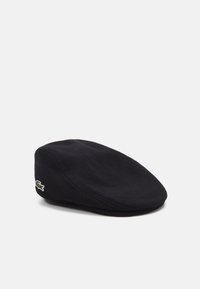 Casquette plate en laine noire avec une couronne arrondie, arborant un petit logo Lacoste vert et blanc sur le côté. Texture douce et design classique.
