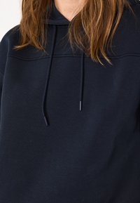 Donna con un hoodie blu scuro con coulisse, capelli sciolti sulle spalle, tagliato a livello del collo e del busto.