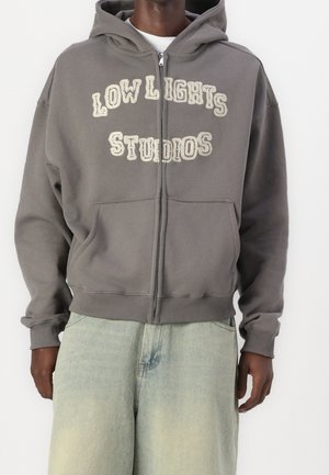 Grå zip-up hoodie med texten "LOW LIGHTS STUDIOS" i krämvit. Har huva, framfickor och ribbade muddar. Mjuk tyg med avslappnad passform.