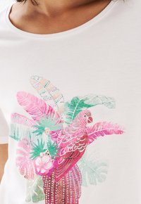 T-shirt blanc avec un imprimé coloré d'un perroquet et de feuilles tropicales en rose, vert et teal, avec des motifs et des textures détaillés.