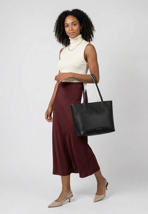 Femme aux cheveux bouclés portant un col roulé sans manches blanc, une jupe midi bordeaux, des talons beiges et un sac fourre-tout en cuir noir.