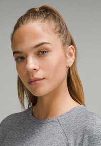 Lähikuva henkilöstä, joka on pukeutunut harmaaseen teksturoituun paitaan. Heillä on pitkät, suorat hiukset ja vasemmassa korvassa pieni kultainen renkaankorvake.