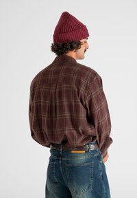 Man met krullend haar, gekleed in een bordeauxrode gebreide muts, een bruine geruite shirt en een spijkerbroek, staand in een driekwart achteraanzicht tegen een witte achtergrond.