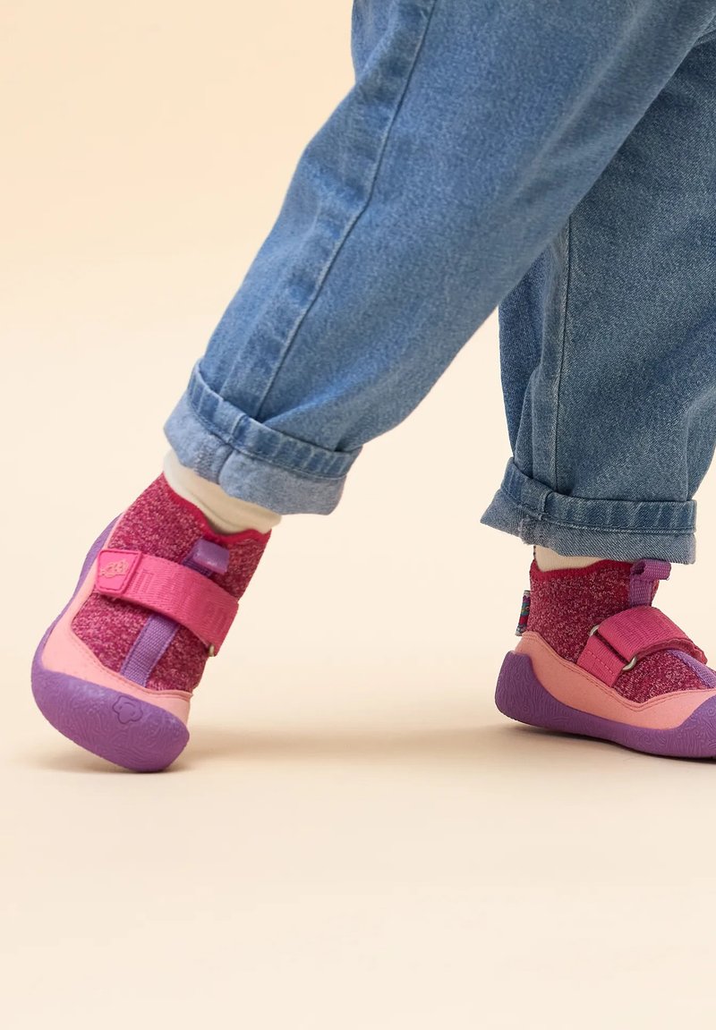 Różowe i fioletowe sneakersy z tkaniny, z regulowanym paskiem i gumową podeszwą, zestawione z niebieskimi spodami dżinsowymi i białymi skarpetkami.
