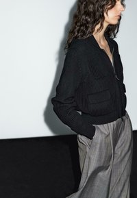 Woman with long dark wavy hair wears a black knit zip-up jacket and high-waisted gray trousers with hands in pockets, facing right.