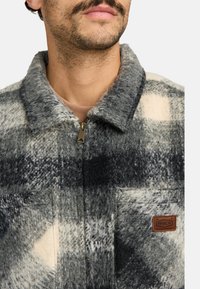 Fleecejacke in grau-creme kariertem Muster mit Reißverschluss, Kragen und einer Brusttasche mit braunem Logo-Patch.