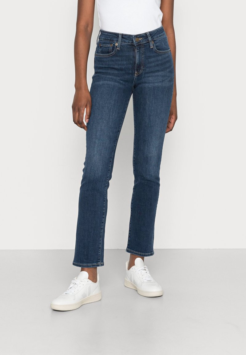 Jeans de mezclilla azul oscuro con corte recto, cintura de altura media, ligeras decoloraciones y diseño de cinco bolsillos, combinados con zapatillas blancas.