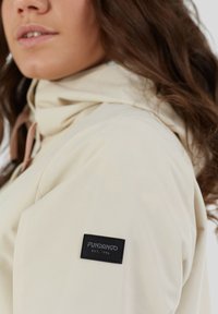 Veste de couleur crème avec une capuche, comportant une étiquette noire sur la manche. La texture lisse et le design minimaliste accentuent son look décontracté.
