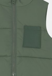 Wyściełana kamizelka bez rękawów w ciemnozielonym kolorze, wyposażona w przedni zamek błyskawiczny oraz pojedynczą kieszeń na piersi, z gładką fakturą i subtelnymi detalami przeszyć.