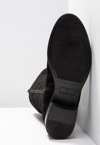 Bottine en daim noir avec une surface lisse et texturée, une semelle en caoutchouc plate, et une fermeture éclair intérieure pour un accès facile.