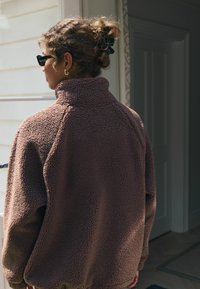 Pullover in pile marrone con colletto alto, superficie testurizzata e spalle scese. Presenta maniche lunghe e una vestibilità rilassata.