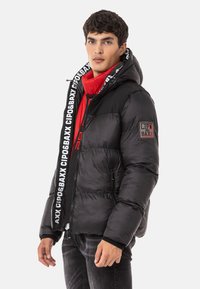 Schwarze, gepolsterte Daunenjacke mit Kapuze, ausgestattet mit einem schwarz-weißen Logostreifen, seitlichen Reißverschlüssen und einem roten Innenfutter. Akzentpatch am Ärmel.
