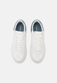 Pár bílých tenisek Calvin Klein s modrou vnitřní výstelkou, bílými tkaničkami a perforovaným jazykem, pohled zeshora na světlém pozadí.