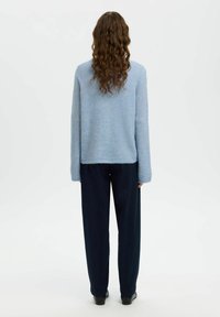 Šviesiai mėlynas megztinis su ilgomis rankovėmis, laisvo silueto, su suraitytu apačios kraštu ir reljefine tekstūra, derinamas su tamsiomis kelnėmis, matomas iš nugaros.