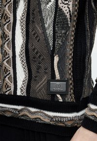 Κοντινή λήψη πλεκτού πουλόβερ με μαύρα, καφέ, μπεζ και λευκά γεωμετρικά σχέδια και ορθογώνια δερμάτινη ετικέτα επωνυμίας στο μπροστινό μέρος.