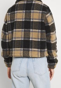 Fleece jacket with a black, tan, and grey plaid pattern. Cropped design, soft texture, and wide sleeves, paired with light blue jeans.