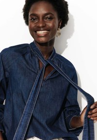 Blouse en denim avec un détail de lien au niveau du col, des manches bouffantes et des coutures contrastantes. Couleur bleu foncé avec une texture lisse.