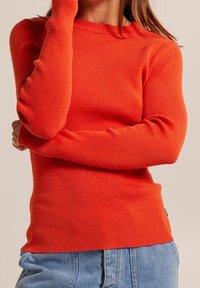 Pull à manches longues côtelé de couleur orange vif, avec un col rond et une coupe ajustée, associé à un jean en denim bleu clair.