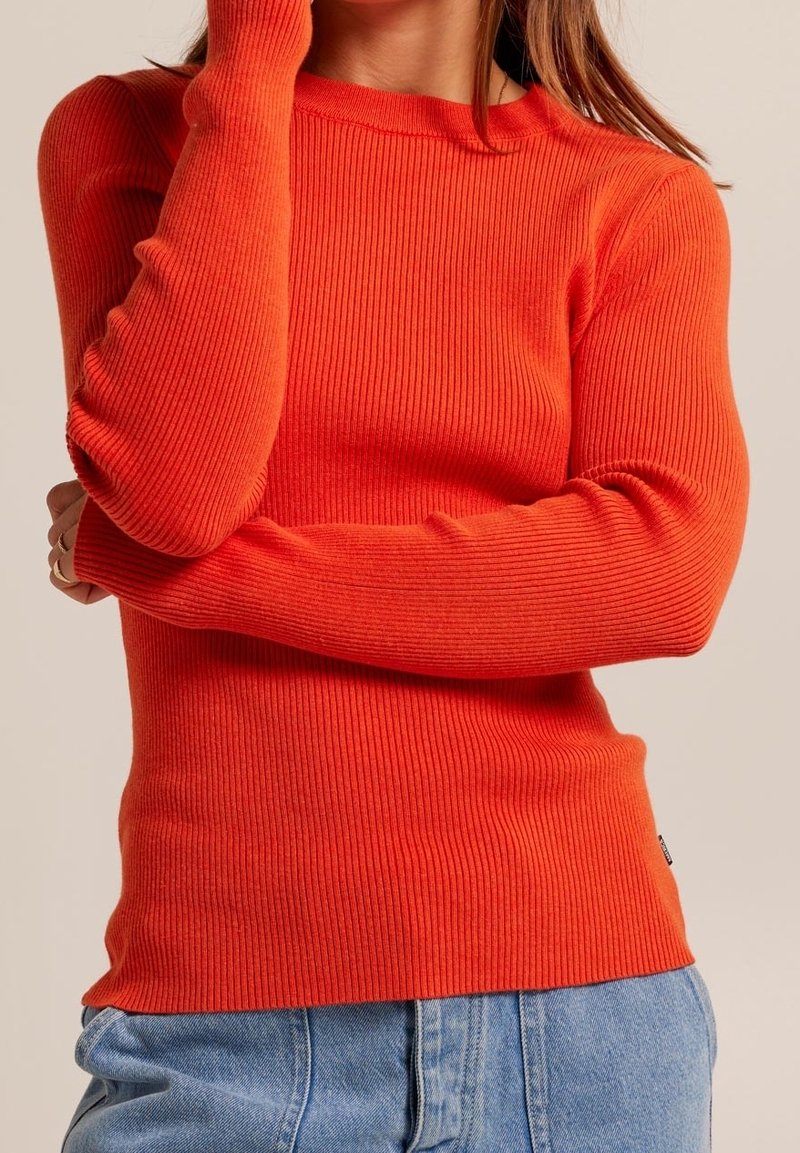Pull à manches longues côtelé de couleur orange vif, avec un col rond et une coupe ajustée, associé à un jean en denim bleu clair.