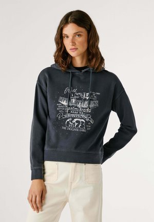 Felpa con cappuccio di un blu navy scuro, realizzata in un mix di cotone, con testo grafico bianco e logo. Presenta un cappuccio con lacci e polsini a costine, abbinata a pantaloni chiari.