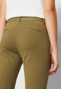 Pantalones verde oliva con una textura suave y elástica, que cuentan con dos bolsillos traseros y un pequeño acento circular de oro rosa.