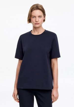Femme aux cheveux blonds courts portant un t-shirt bleu marine uni à manches courtes et un pantalon assorti sur fond blanc.