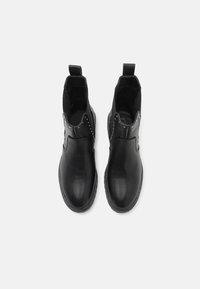 Paire de bottines en cuir noir avec languettes et bord clouté autour de la cheville sur fond blanc.