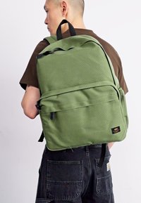 Zaino verde realizzato in tessuto texturizzato, con tasca anteriore con cerniera, manico superiore e spallacci neri, indossato da un individuo.