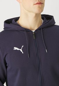 Námořnická modrá mikina na zip vyrobená z bavlněné směsi, s bílým logem Puma na levé hrudi a nastavitelnými stahovacími šňůrkami na kapuci.