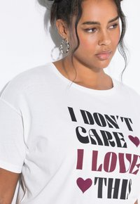 Mujer con camiseta blanca que lleva texto negro y burdeos "NO ME IMPORTA, ME ENCANTA ESTO" y gráficos de corazones en burdeos, pendientes de aro plateados, expresión neutral.