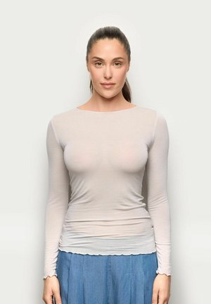 Młoda kobieta o ciemnych włosach związanych do tyłu, ubrana w dopasowaną prześwitującą beżową bluzkę z długim rękawem oraz niebieską spódnicę dżinsową, zwrócona przodem na jednolitym tle.