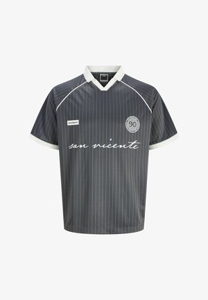 Ung person med krøllet hår iført sorte solbriller, mørk stribet kortærmet skjorte, lyse denimshorts, hvide sokker og brune støvler.