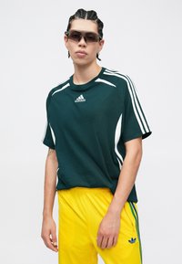 Ciemnozielona koszulka z krótkim rękawem z białymi bocznymi paskami i logo Adidas, zestawiona z jasnżółtymi spodniami z bocznymi paskami i logo.