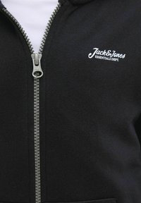 Čierna mikina na zips vyrobená z mäkkej tkaniny. Má centrálny zips, rebrovanú textúru a biele logo "Jack & Jones Essentials Dept." na hrudi.