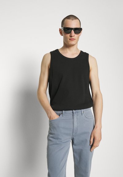 Black sleeveless shirt in a ribbed texture, paired with light blue denim jeans. Model wearing black sunglasses, hands in pockets.