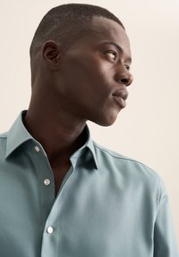 Chemise bleu clair à manches longues avec un col net, un tissu lisse et des boutons blancs. Détails de couture subtils le long des coutures des épaules.