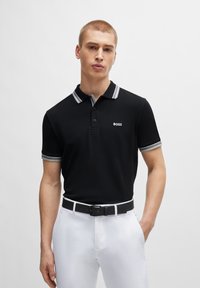 Polo noir avec un col et des poignets rayés gris contrastants, présentant une patte de boutonnage à l'avant et un petit logo "BOSS". Associé à un pantalon blanc.