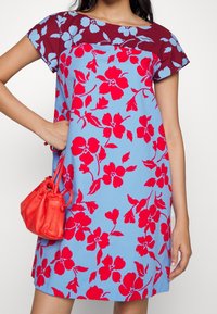 Květinové šaty s modrou základnou a červenými květy. Tmavě červený panel na výstřihu. Krátké rukávy a volný střih. Součástí je malá červená kabelka.