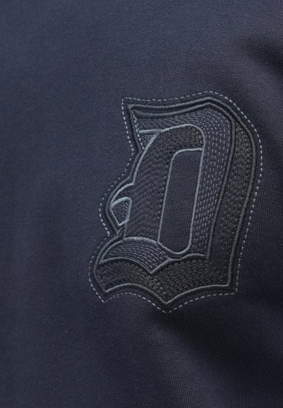 Marineblå stof med en tekstureret lap, der har et sort broderet bogstav "D" og kontrasterende sømdetaljer.