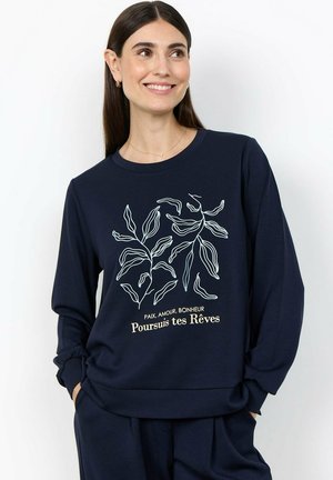 Vrouw met lang donker haar glimlacht, draagt een marineblauwe sweatshirt met een wit botanisch ontwerp en Franse tekst "Poursuis tes Rêves."