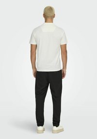 T-shirt blanche à manches courtes avec accent au dos, associée à un pantalon noir ample et des baskets beiges, montrant un design décontracté.