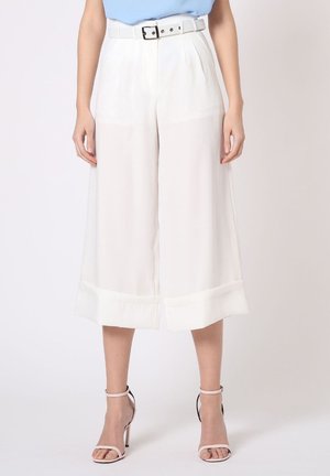Mujer con pantalones blancos de pierna ancha y cortos con cinturón y sandalias de tacón alto color rosa claro, de pie sobre un fondo liso.