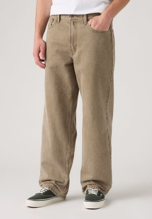 Personne portant un jean ample marron clair et des baskets basses noires avec des semelles et lacets blancs, debout devant un fond uni.
