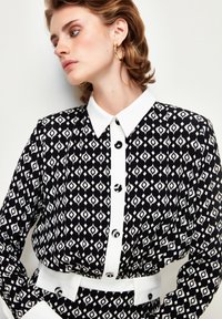 Black and white patterned blouse with a collared neck, button front, and white belt. Features geometric designs and glossy black buttons.