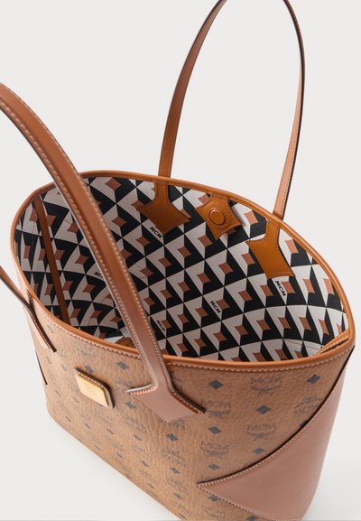 Καφέ δερμάτινη τσάντα tote με μοτίβο λογότυπου MCM, διαθέτει λουριά από δέρμα σε χρώμα ταμπά και εσωτερικό υφασμάτινο με γεωμετρικό σχέδιο σε μαύρο, λευκό και καφέ.